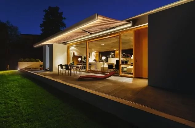 Modernes Einfamilienhaus mit beleuchteter Terrasse und ausgefahrener Markise am Abend – hochwertige Markisenlösung für Terrassen in Braak.